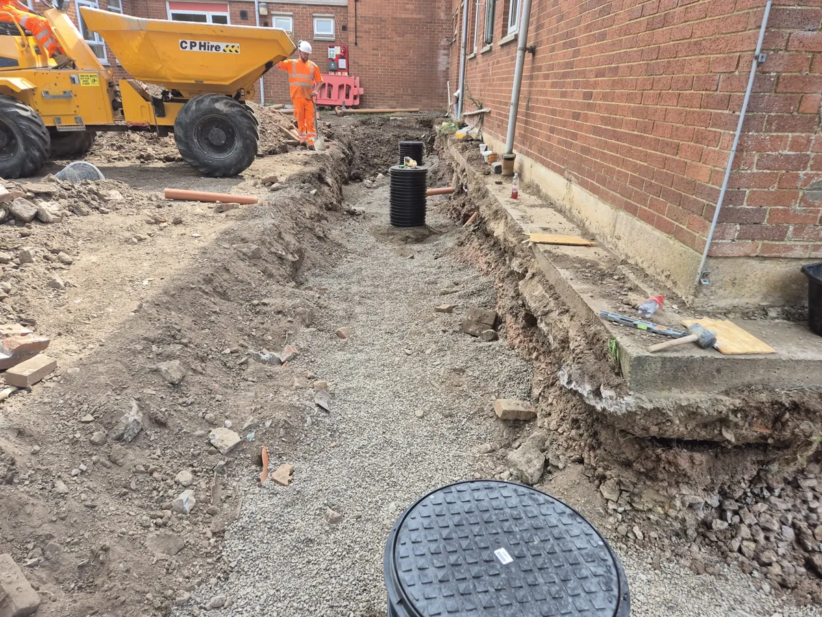 A drainage trench and inspection chamber dug alongside a building, with a dumper on site