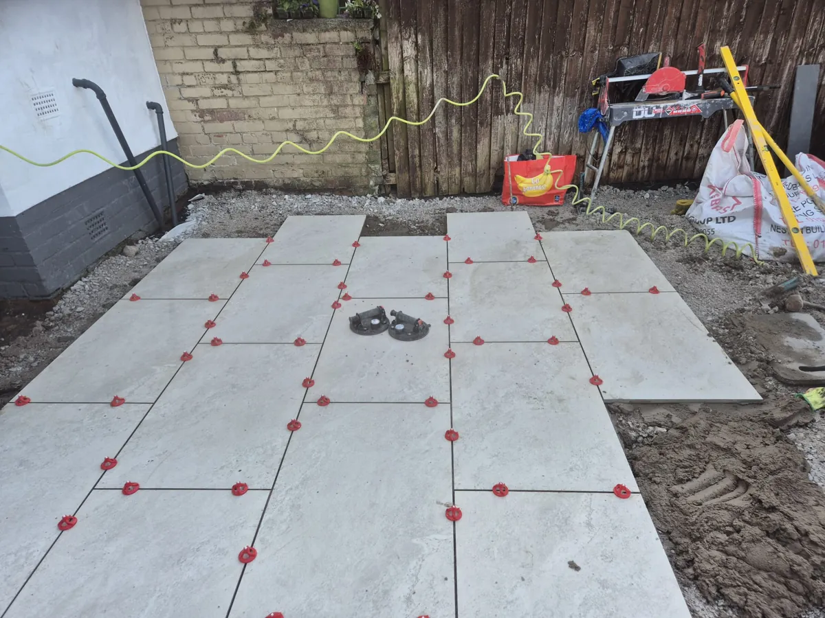 Most of a light grey porcelain patio laid on a full mortar bed in Birkenhead, with levelling clips in place and a tile saw set up against the fence